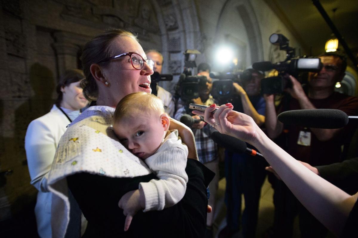Karina Gould speaks to media while holding a small child, surrounded by journalists and photographers in an indoor setting at night.